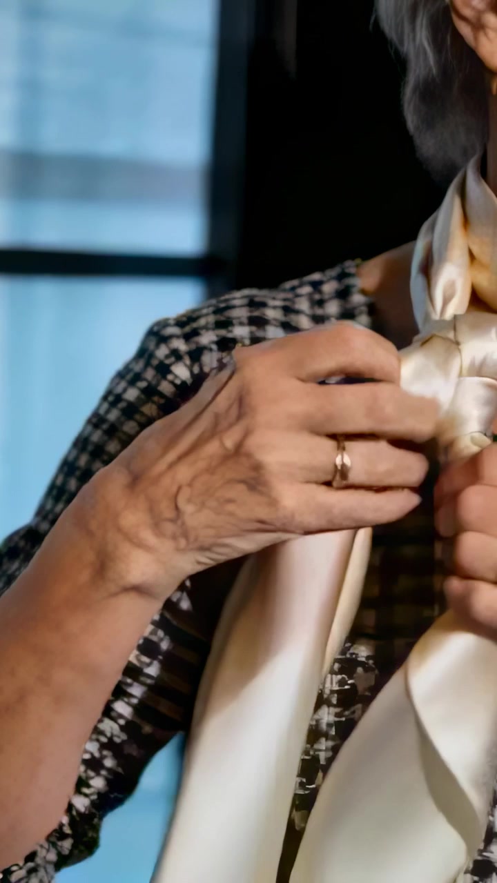 Video verticale: Donna che sistema l'estremità sinistra e il nodo di una sciarpa color crema vicino alla finestra, preparando l'outfit