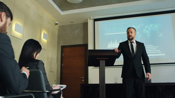 Camera zoom in out on Caucasian businessman talking on a podium in a conference room and showing some charts and graphics on the big screen