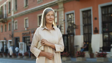 Portrait-Shot-Of-Old-Beautiful-Woman-With-Purse-Posing-To-Camera-And-Smiling-At-Street-In-City-Center