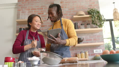 Happy-diverse-couple-in-aprons-using-tablet-and-baking-in-kitchen,-copy-space,-slow-motion