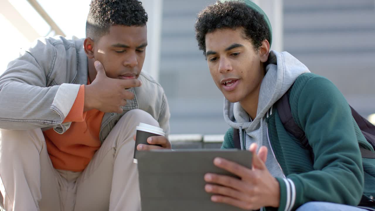 Diverse male friends pulling tablet from bag, swiping, studying on school steps with coffee