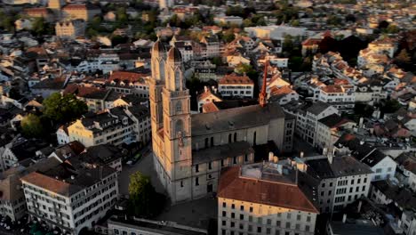 Spinning-aerial-view-of-a-glowing-Grossmünster-church-at-sunset-in-Zurich,-Switzerland,-fast