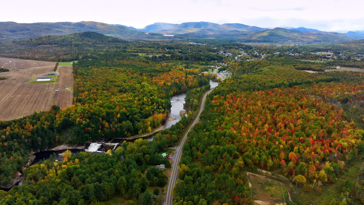 Colorful woods crossed by the narrow river and highway going along it. Mountains at backdrop. Aerial view.