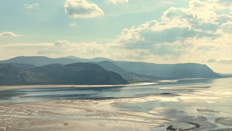 Langsam-Steigendes-Wasser-Nach-Ebbe-Im-Tidebereich-Vor-Der-Küste-Von-Wales-Mit-Den-Bergen-Der-Gebirgslandschaft-Im-Hintergrund-Von-Der-Sonne-Zwischen-Den-Wolken-Beleuchtet