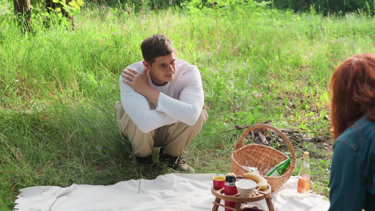 Mother and son engage in gentle conversation during picnic in sunny meadow, surrounded by lush grass, as she prepares to drop used tissue, creating calm and natural family interaction moment