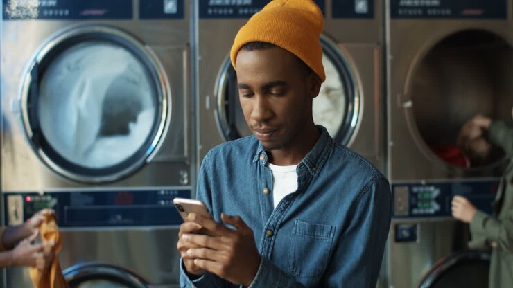 Stylish Man In Yellow Hat Tapping And Texting Message On Smarphone While Sitting In Laundry Service Room