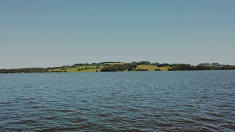 Aerial:-Low-flying-drone-skimming-the-water's-surface-as-it-fly's-at-speed-over-a-large-lake-towards-rolling-green-hills-on-the-other-side