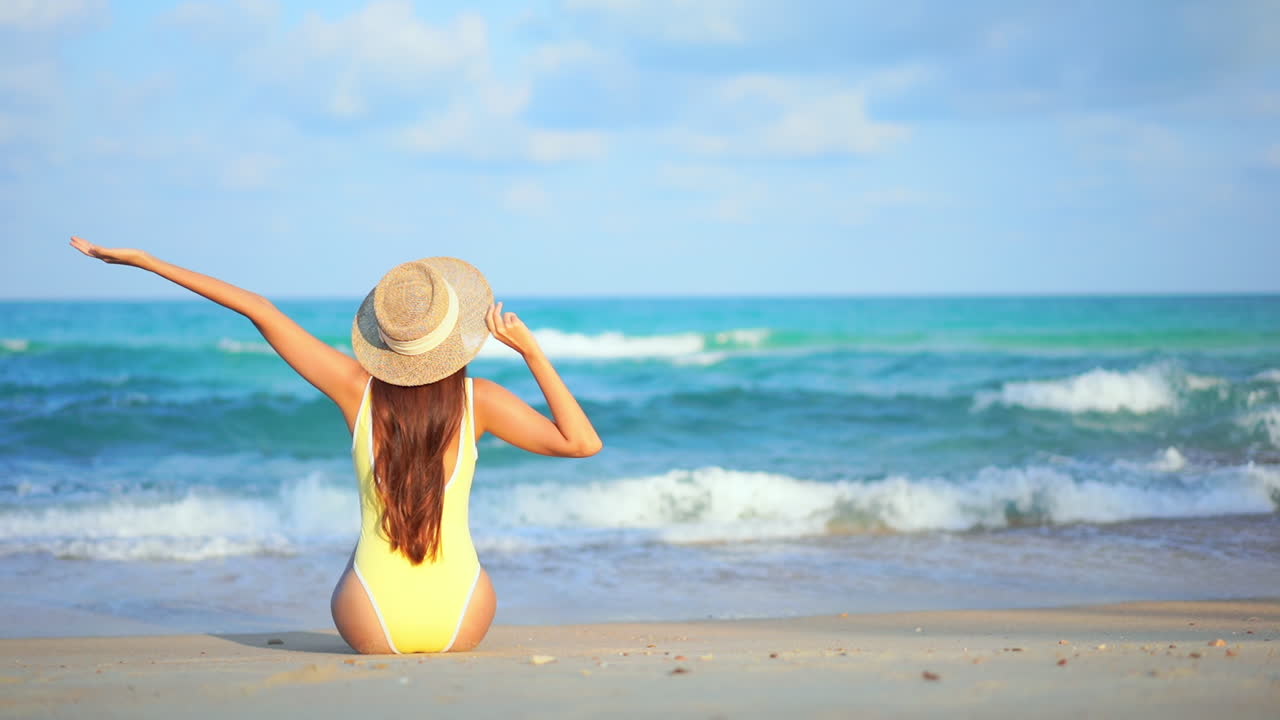 카메라에 등을 대고 해변에 앉아 원피스 수영복을 입은 여성이 뻗은 팔로 들어오는 파도를 맞이합니다.