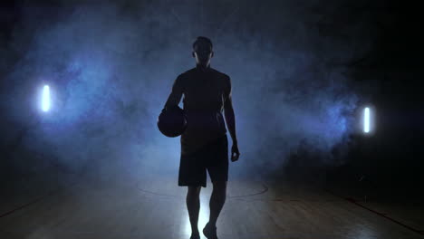 Männlicher-Basketballspieler,-Der-Sich-Im-Rauch-Vor-Der-Kamera-Bewegt-Und-Den-Ball-In-Zeitlupe-Mit-Steadicam-Aufnahme-über-Den-Parkettboden-Wirft