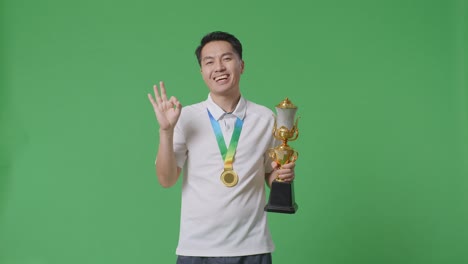 asian man with a gold medal and trophy showing okay gesture and smiling to camera as the first winner on green screen background in the studio