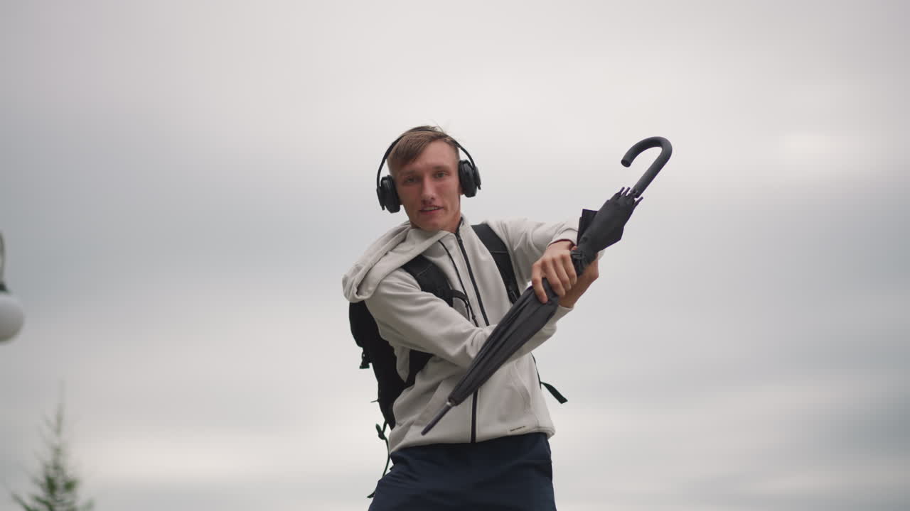 Man with umbrella performing playfully, Young man dancing with umbrella during windy weather, Young individual executing lively umbrella movements on breezy city street in overcast weather