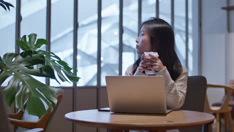 Nostalgic-young,-pretty-Asian-woman-sits-in-cafe-in-front-of-her-laptop,-holding-a-up-of-coffee,-thinking-about-life