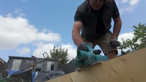 low angle, wide shot of a man planing a plank of wood in garden in 4 x slow motion on a sunny day.