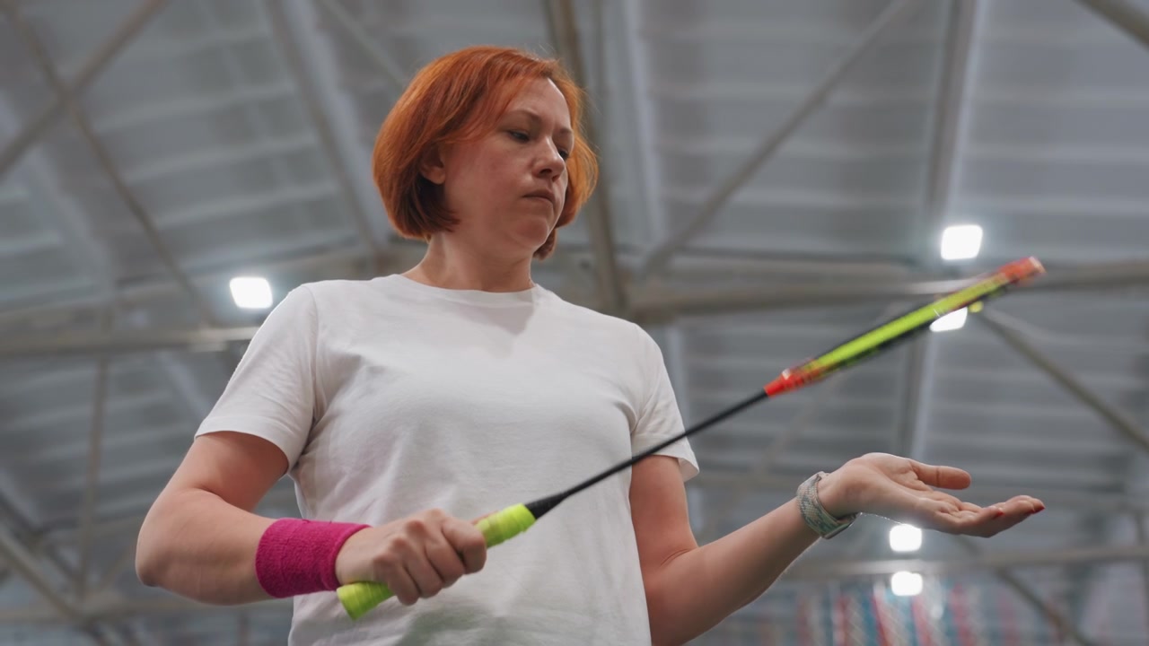 Allenamento di badminton indoor, donna bianca che ispeziona le corde della racchetta, primo piano, inquadratura dal basso che mostra il polsino, luci della palestra, espressione concentrata, mentre regola la presa durante la routine pre-partita, impianto sportivo