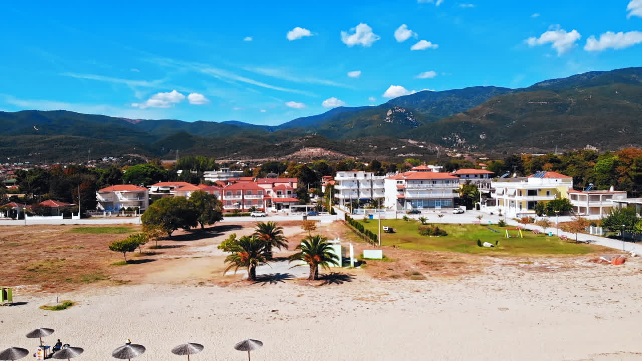 Panorama of the Asprovalta with multiple buildings and greenery, green hills on the background. Aegean sea coast. Beach with umbrellas and sunbeds. Sunny day. Greece