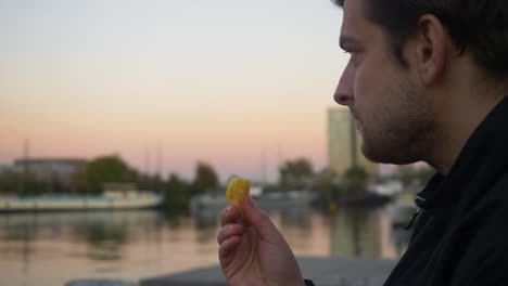 close up of a young man taking an orange slice and eating it on a pier during sunset