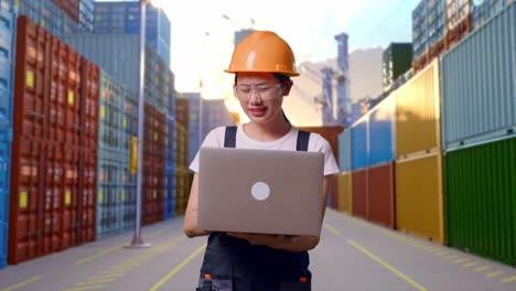 trabajadora asiática con gafas de protección y casco de seguridad usando una computadora portátil y con dolor de cabeza mientras está de pie en un almacén de contenedores