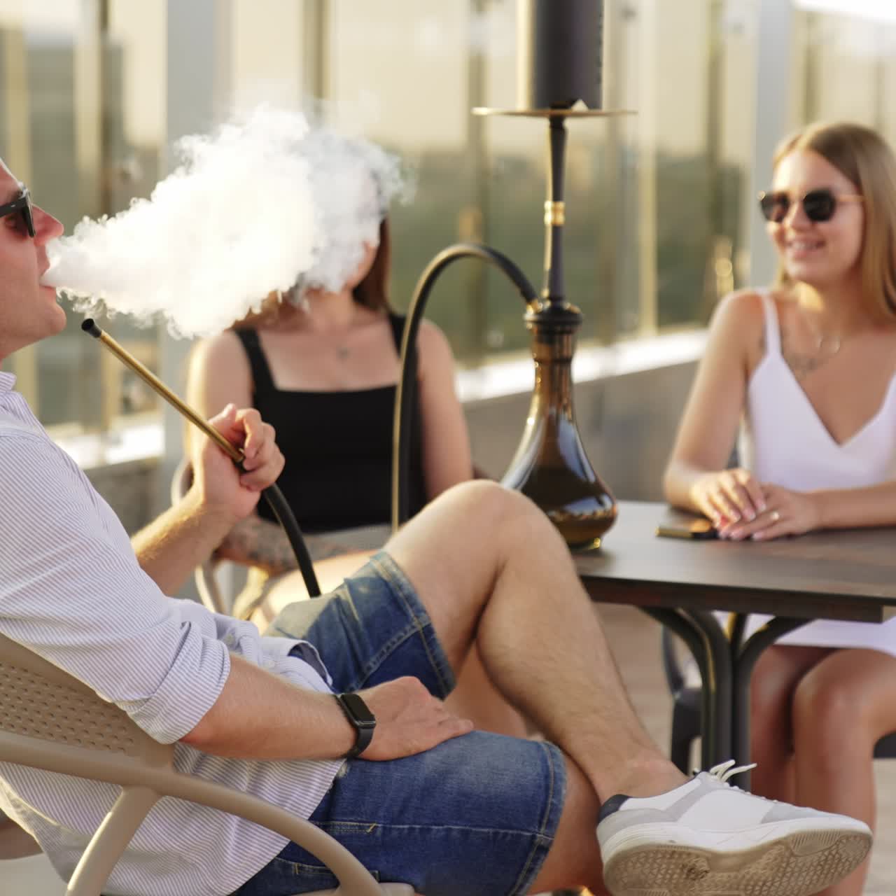Relaxed man sitting in the company of two girls smoking hookah. Young people are chatting enjoying time in a café