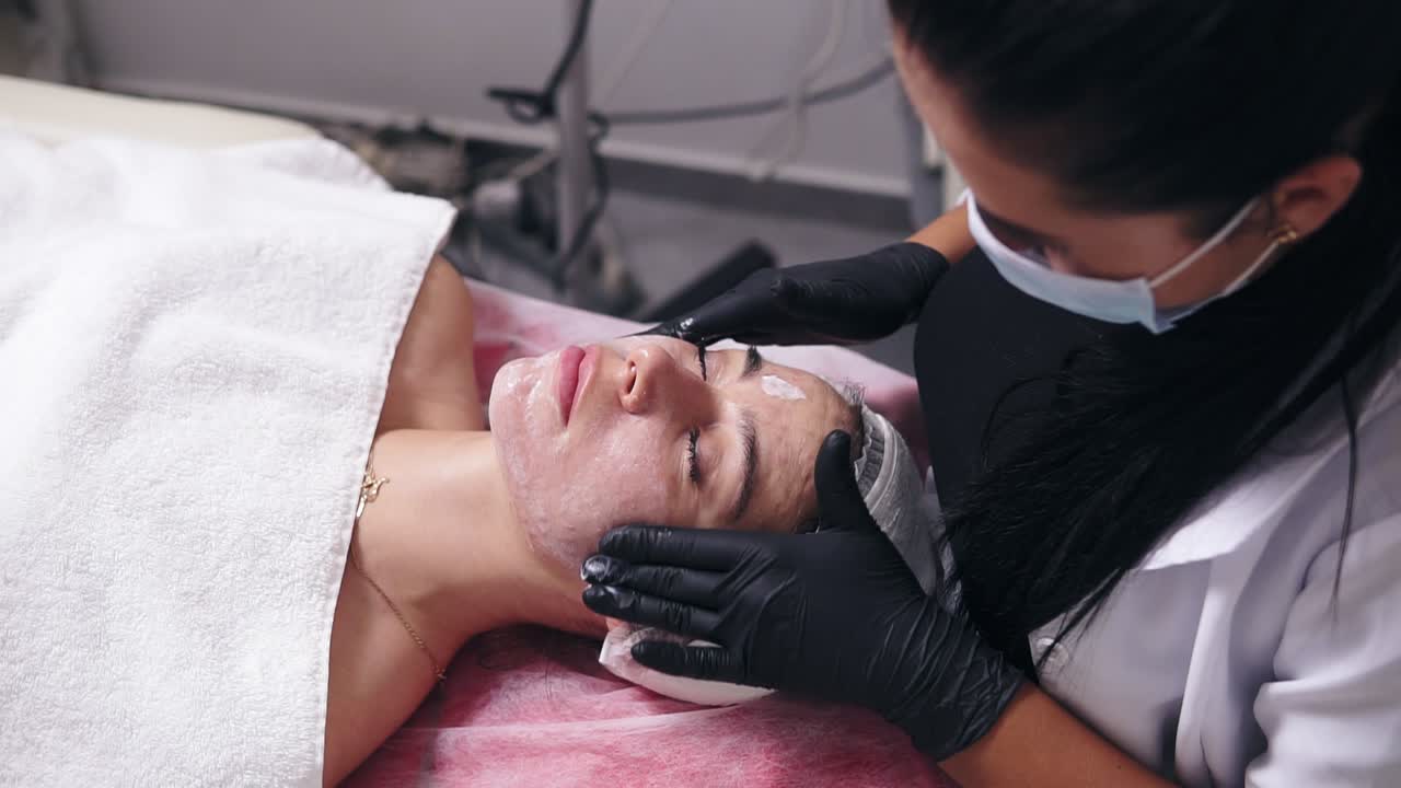 Young woman with blemish skin lying on the couch while professional cosmetologist is applying face cream and making face massage