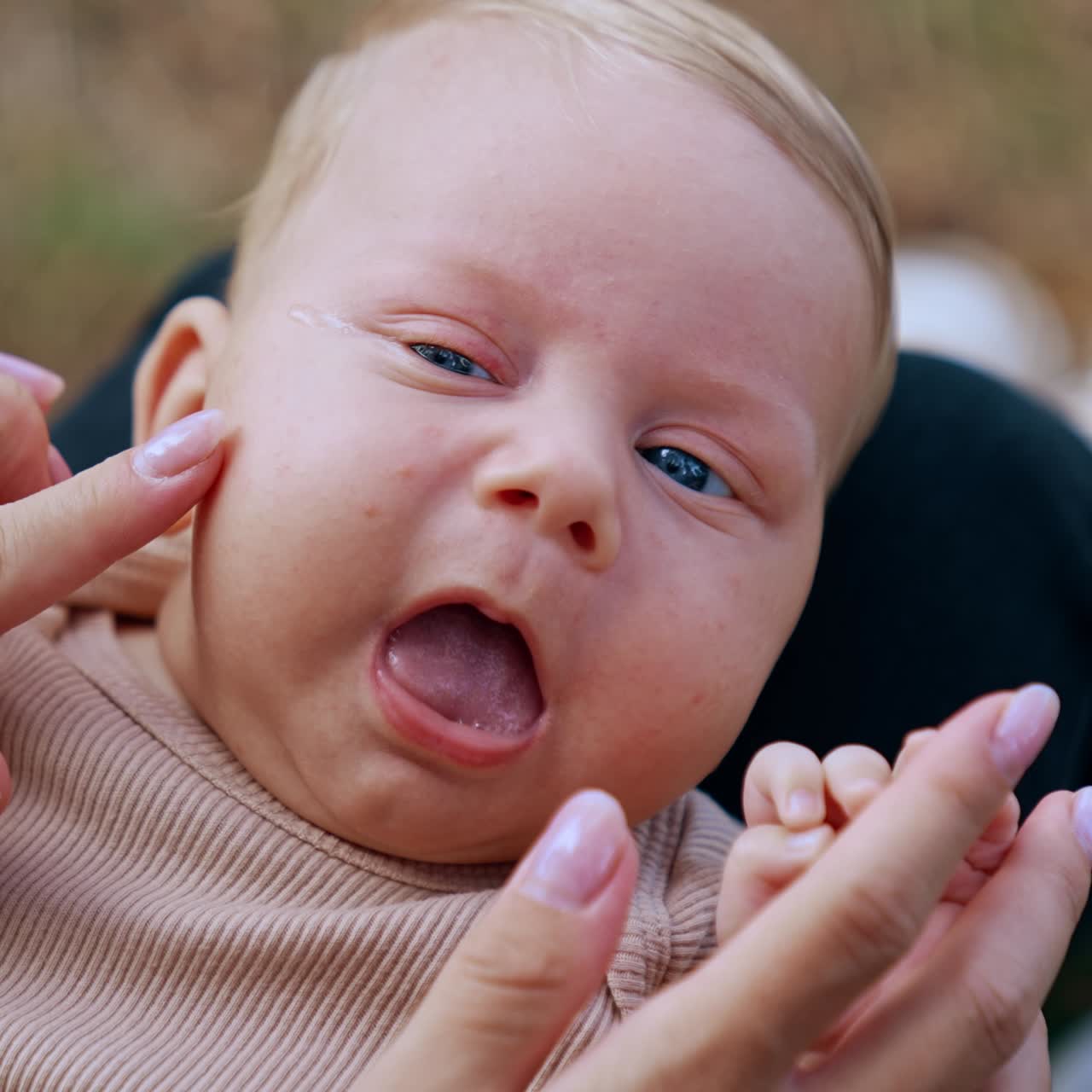 Blond baby boy with adorable blue eyes lies on mom's laps. Mother touches her kid's cheeks and nose. Close up