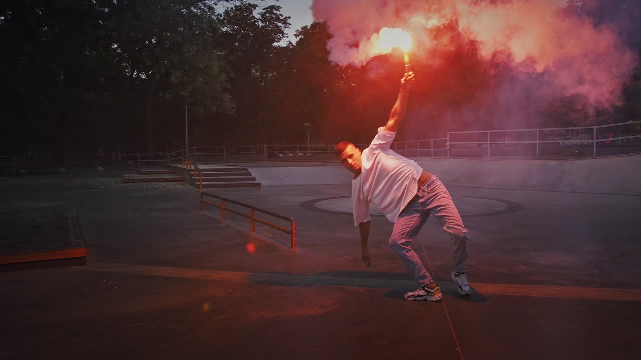 joven macho está sosteniendo una llamarada de señal roja brillante y de pie en postura mientras realiza break dance en la pista de bombeo del rollerdrome. cámara lenta