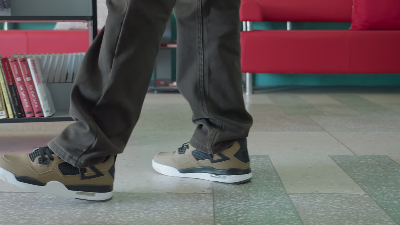 Low angle close up of adult wearing brown pants and beige sneakers walking past bookshelves filled with colorful books in study room, legs in motion on tiled floor with turquoise wall background