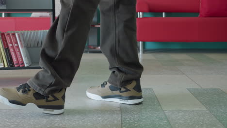 low angle close up of adult wearing brown pants and beige sneakers walking past bookshelves filled with colorful books in study room, legs in motion on tiled floor with turquoise wall background