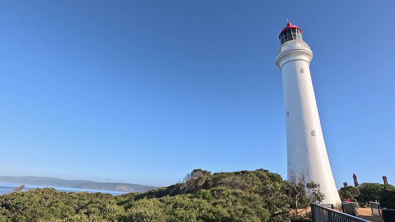 Un alto faro bianco con tetto rosso si erge sopra la macchia costiera in una giornata di cielo sereno