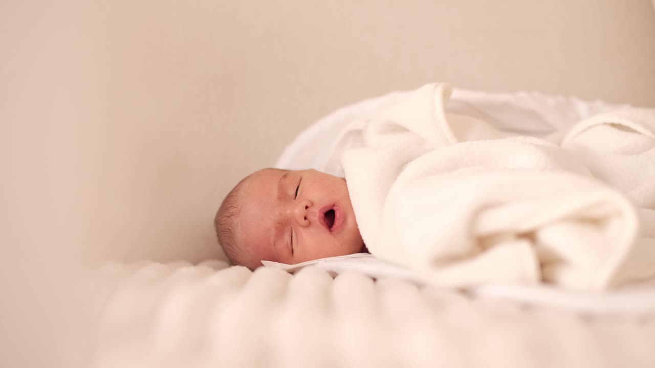 Adorabile neonato sdraiato su un morbido divano bianco, coperto da una coperta, che si addormenta lentamente.