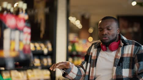 A-happy-man-with-Black-skin-color-in-a-checkered-shirt-and-in-red-wireless-headphones-dances-among-the-counters-in-a-modern-grocery-store-and-has-fun-during-his-shopping