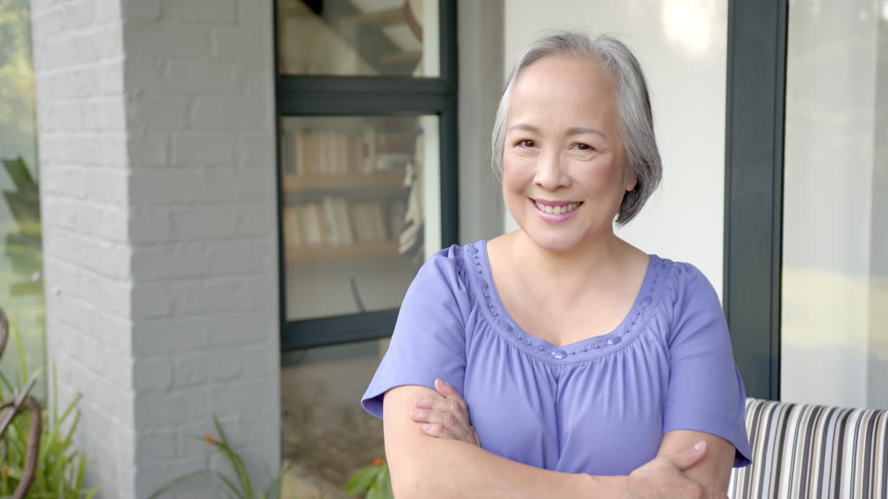 Smiling senior asian woman standing with arms crossed outside home, looking confident