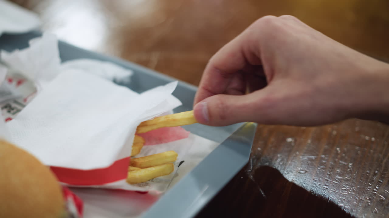 White man hand reaching for fry from container while phone rests nearby on table, closeup showing fingers grasping crispy stick, captured with soft background blur and light glint on surface