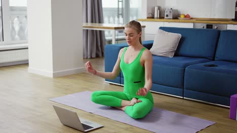 Mujer-En-Ropa-Deportiva-Habla-En-Videollamada-Sentada-En-Una-Estera-De-Yoga-Saluda-En-Namaste