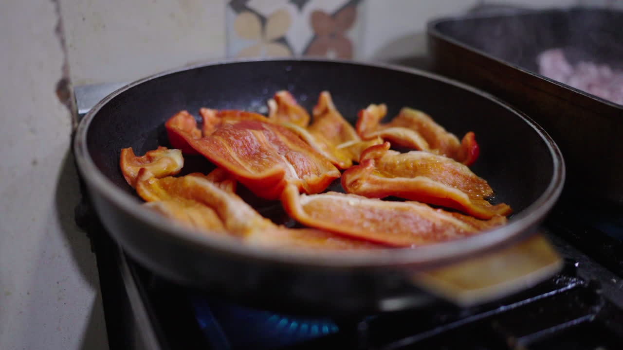 Close-up view during frying some sliced red bell peppers in a black pan over a blue flame stove.