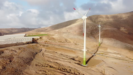 Fantastic-aerial-shot-in-orbit-of-two-wind-turbines-and-a-large-greenhouse-in-a-desert-landscape-and-during-sunset