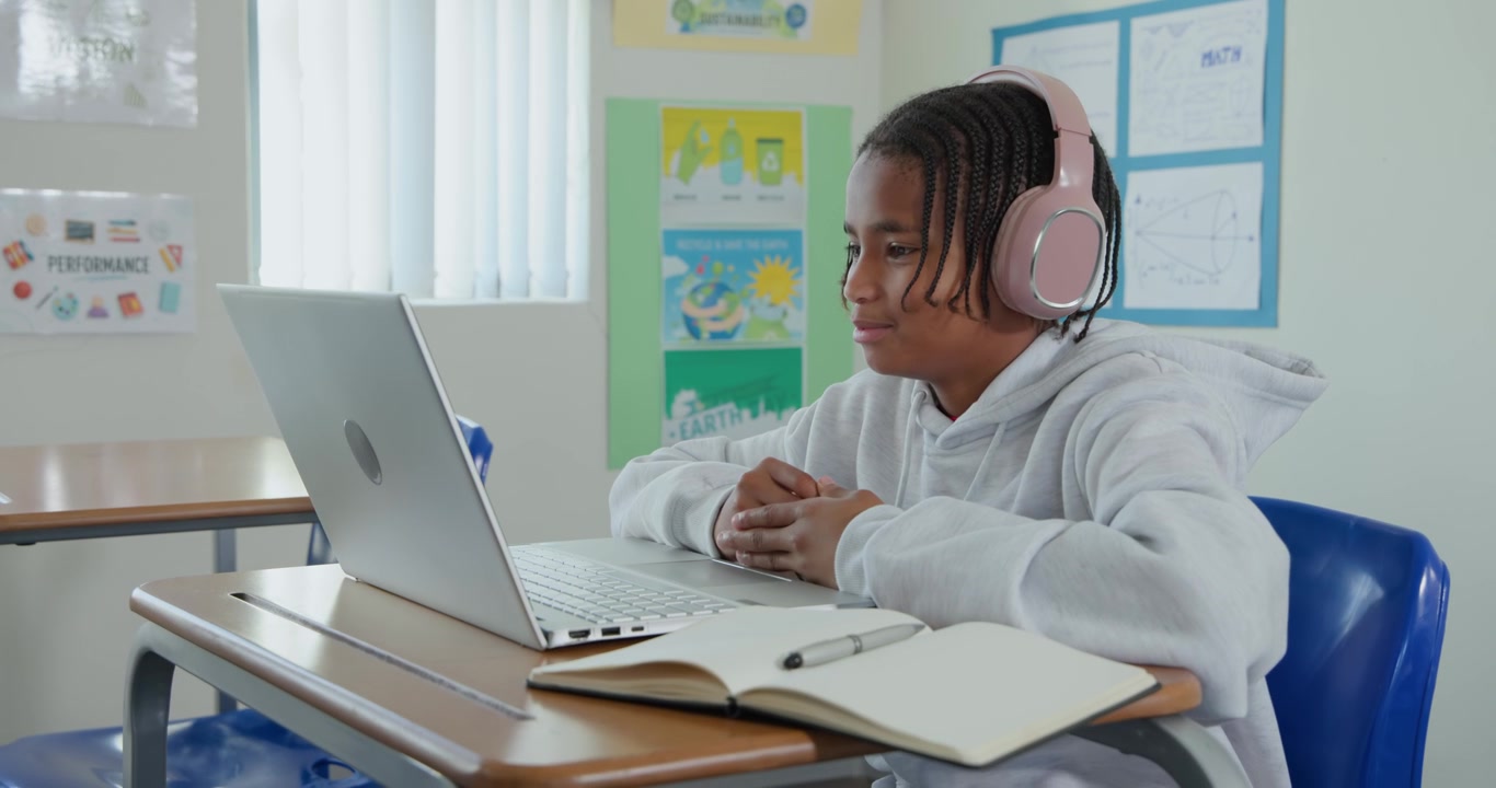 Adolescente afro-americano participando de aula no laptop na escola, usando fones de ouvido rosa, falando.