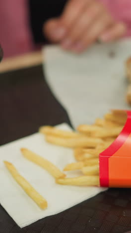primer plano de la mano de una mujer joven en vestido rosa extiende la mano por las papas fritas de una bandeja, con una hamburguesa, papas fritas y taza de café cerca de una servilleta y comió tomó el café también y bebió de él