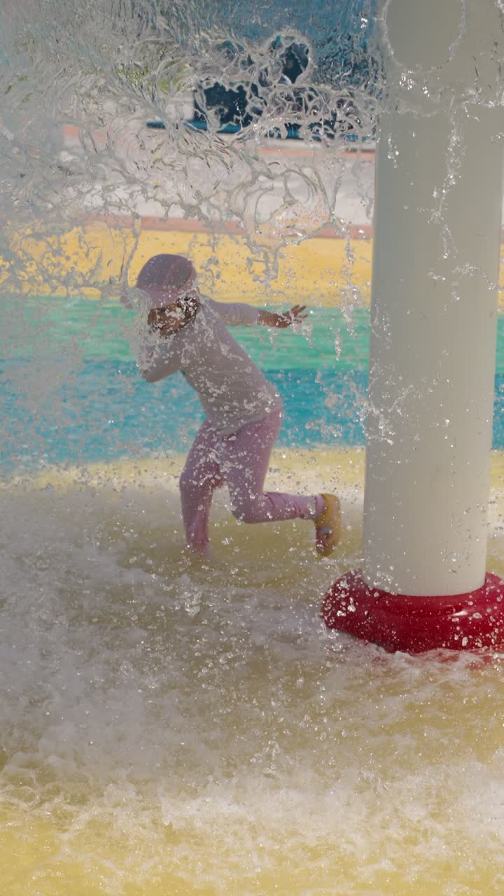 High-speed 120fps slow motion vertical shot of a five-year-old girl splashing and playing with water jets at a vibrant outdoor public water playground on a summer day
