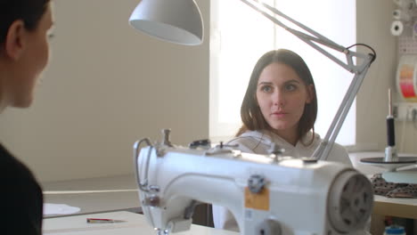 young woman is working with sewing machine and checking stitches when her colleague is coming to her with sketch. women. are looking at fabric and talking