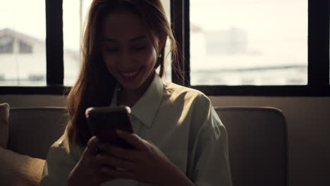 a woman browses the internet using her smartphone while sitting on a couch, smiling as she reads through her social media.