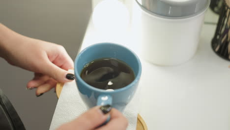 beautician gently holds blue ceramic tea cup on small wooden tray with white tissue, showcasing black nail polish and precise care while serving warm beverage in cozy, professional salon setting