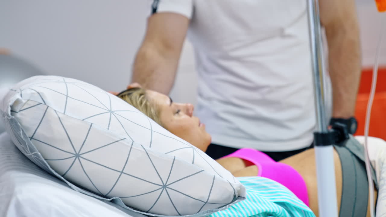 Loving man stands at the bed of his pregnant wife. Blonde lady breathing deep to relief painful contractions.