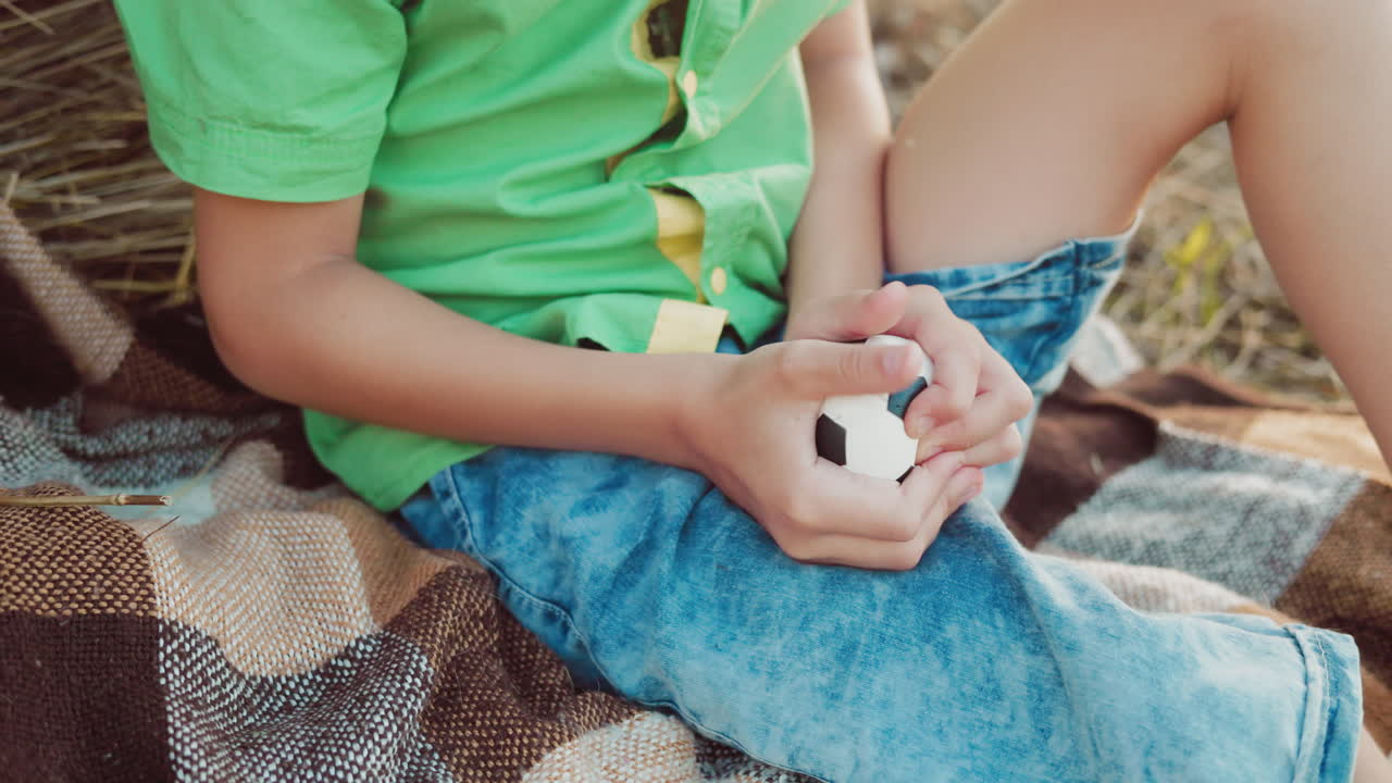 vista parcial de un niño pequeño en vaqueros azules y una tapa verde presionando una pelota de juguete negra y blanca, sentado al aire libre cerca de una bola de heno durante un día soleado