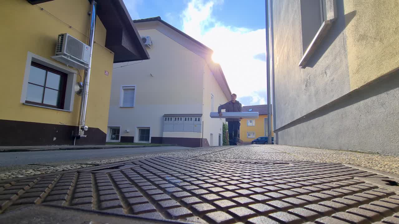 A man in workwear carries a heavy cardboard box or furniture component through an alleyway between houses during a bright day