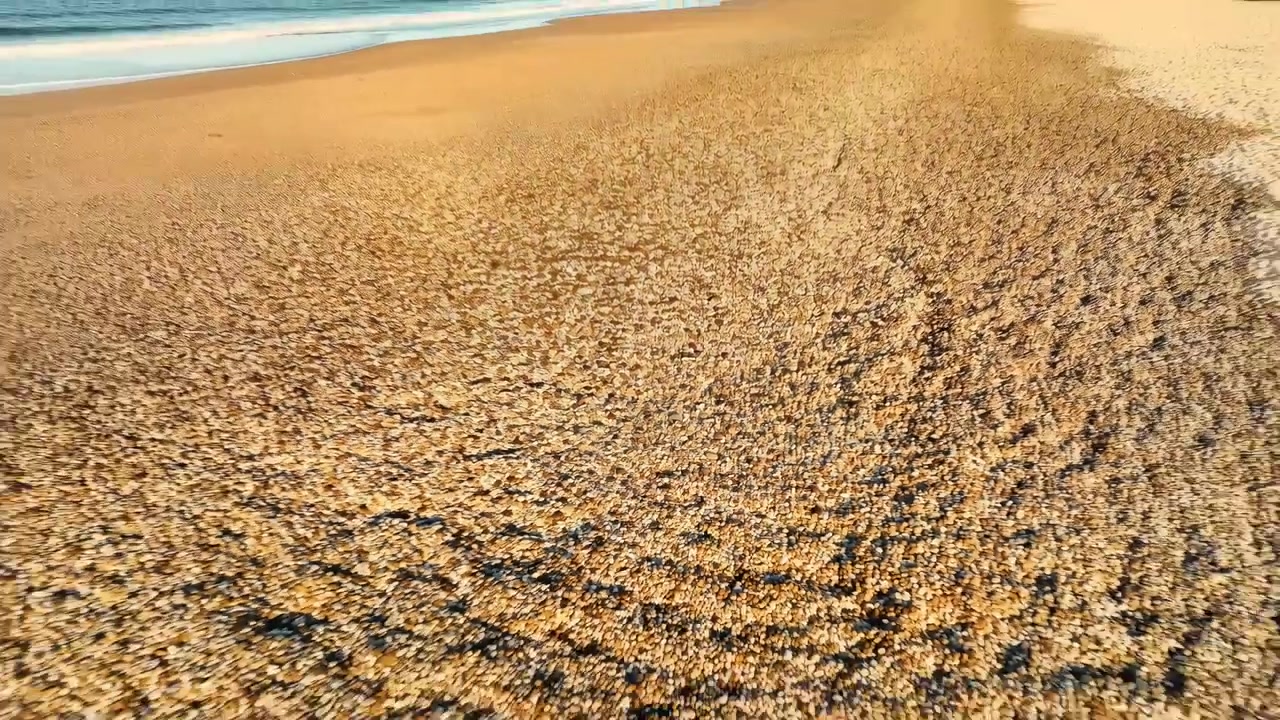 Vista de baixo ângulo varrendo uma costa coberta de seixos, revelando uma transição gradual de pedras arredondadas para areia clara e arrebentação rasa, com tons quentes de pôr do sol, superfície texturizada e paisagem costeira calma.