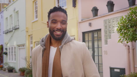 portrait of smiling man wearing coat and scarf standing on cobbled mews street on visit to city in autumn or winter - shot in slow motion