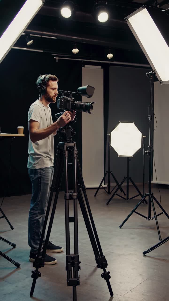 A videographer adjusts a camera on a tripod in a studio. The side angle captures the setup