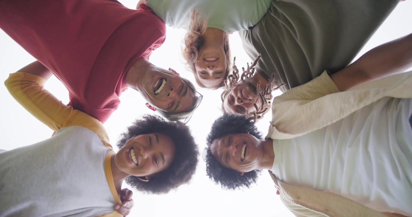 Amigos diversos interagindo com a câmera no chão, inclinando-se enquanto capturam uma tomada de ângulo baixo, rindo ao ar livre.