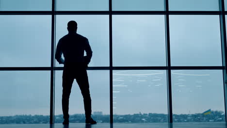sportive male silhouette walks along the panoramic window in office. man holds the laptop and glasses in his hands. low angle view.