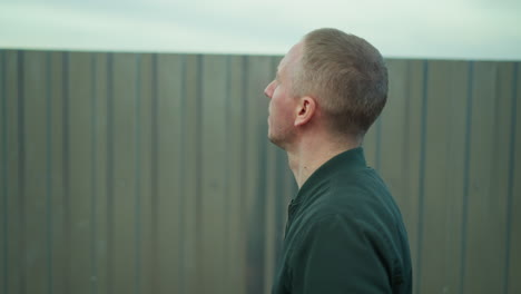 a close view of a man in a green jacket walking alongside a zinc fence, looking around the surrounding